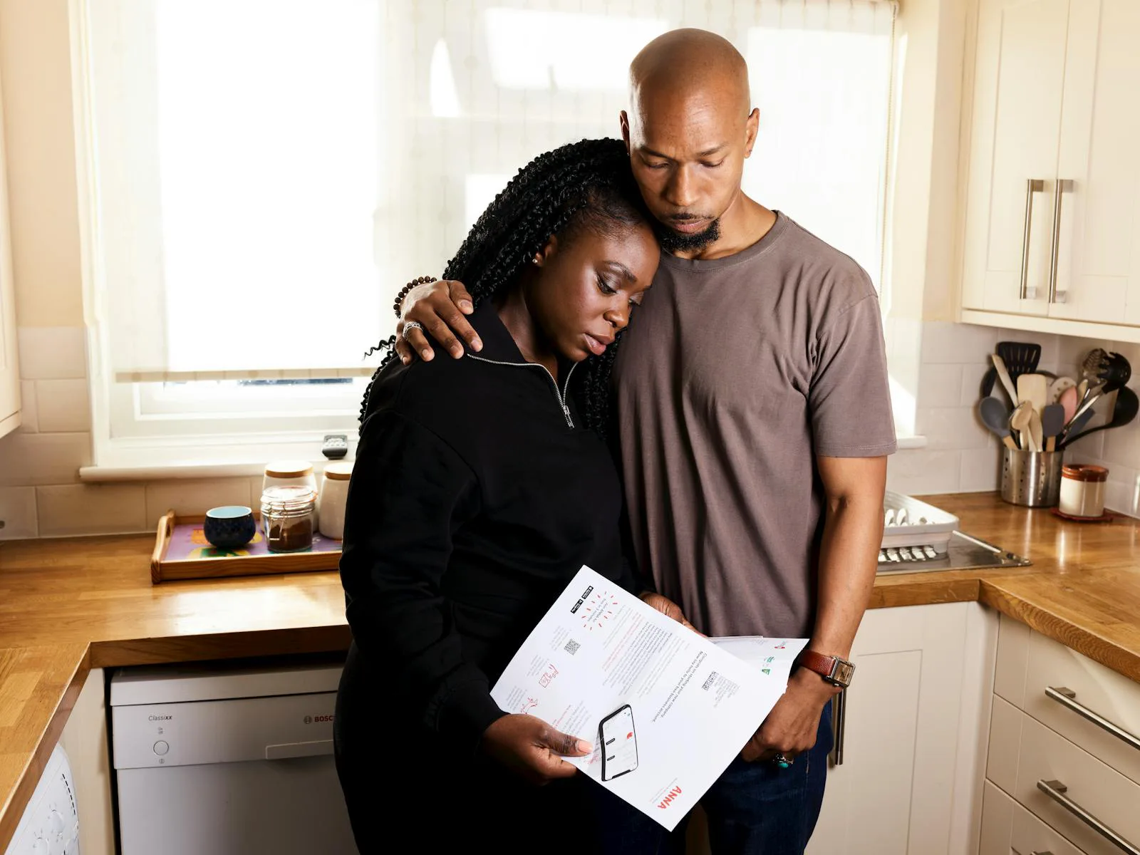 A couple in a sunlit kitchen, leaning into each other while reading a document together.