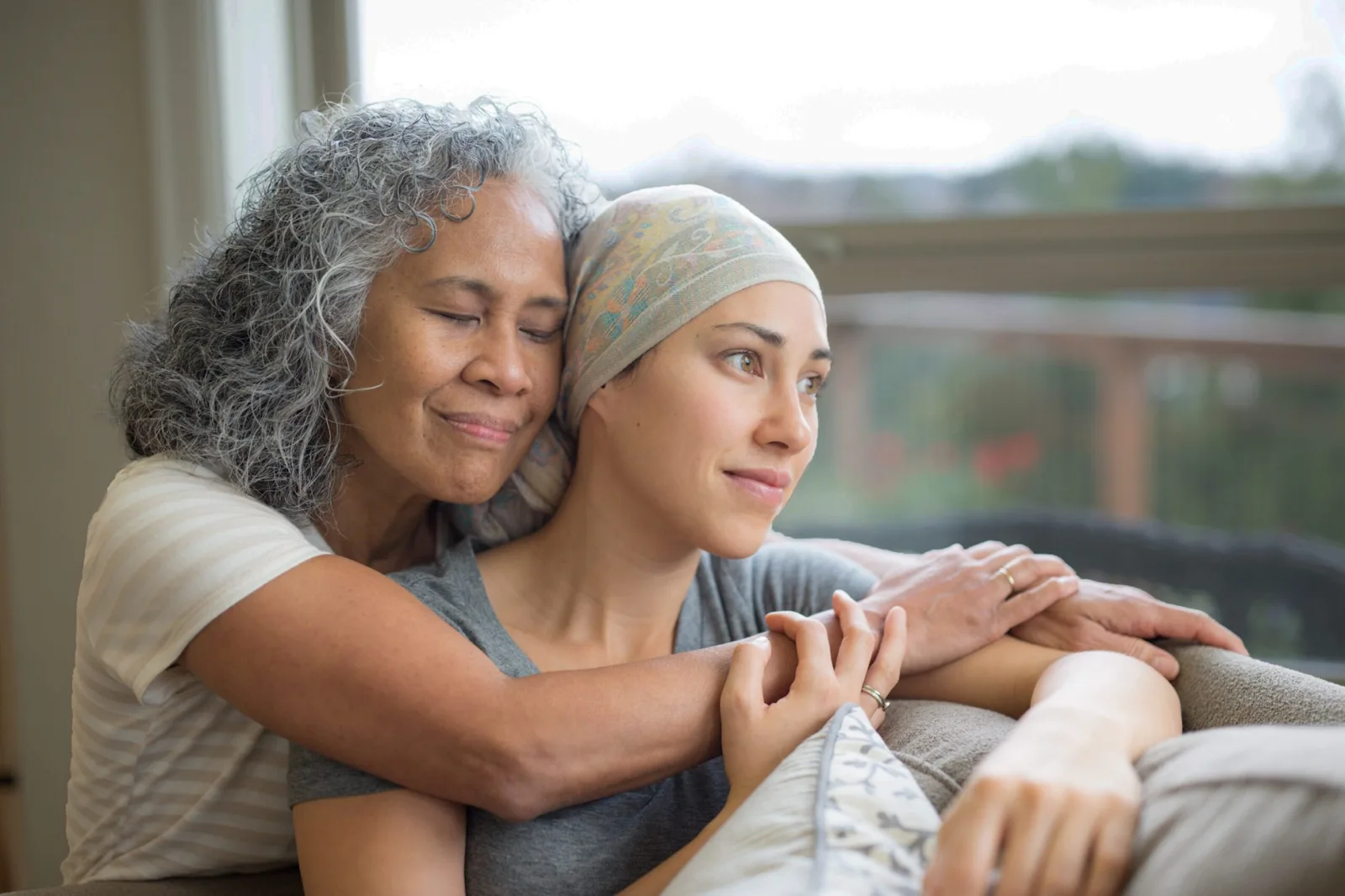 An older woman holding a younger woman in a headscarf, both looking out a window.