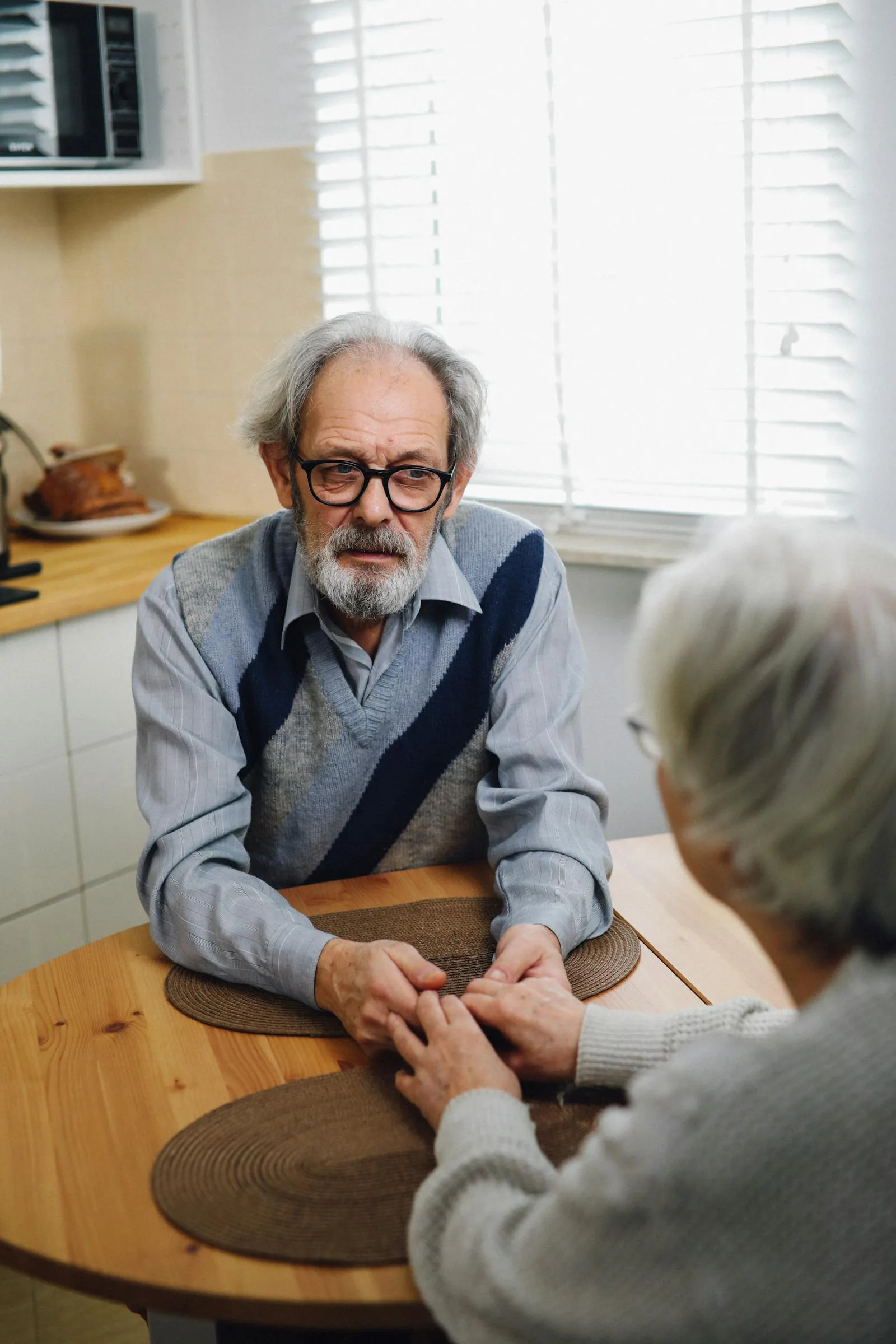 An older couple sitting at a kitchen table holding hands during a serious conversation.